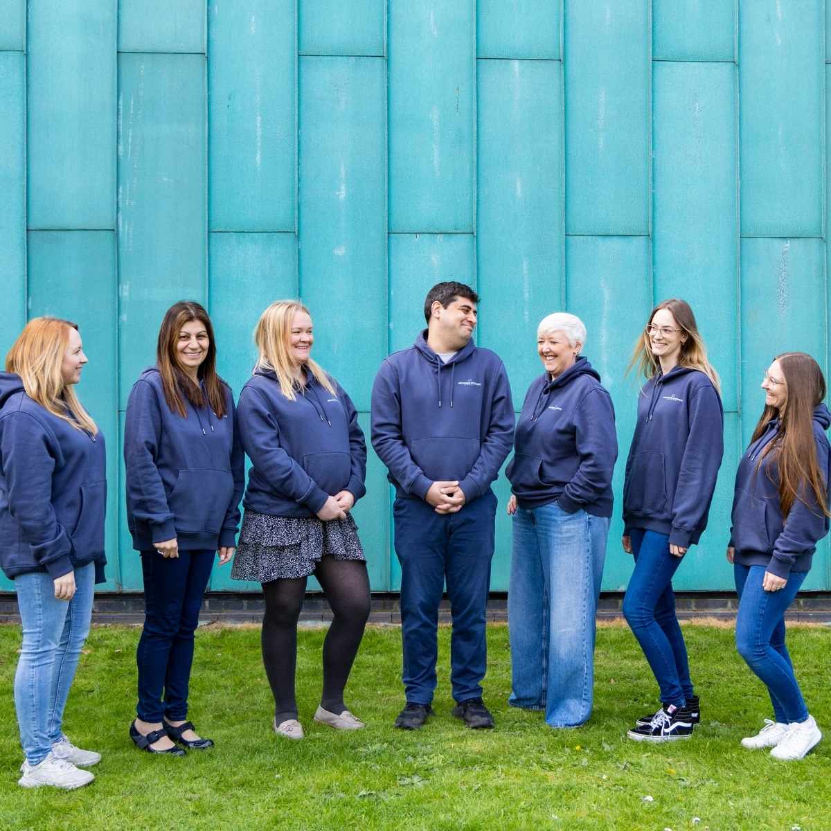 Team Photos - Archimedia Accounts - full team shot of everyone wearing branded hoodies, stood in front of a bright blue wall