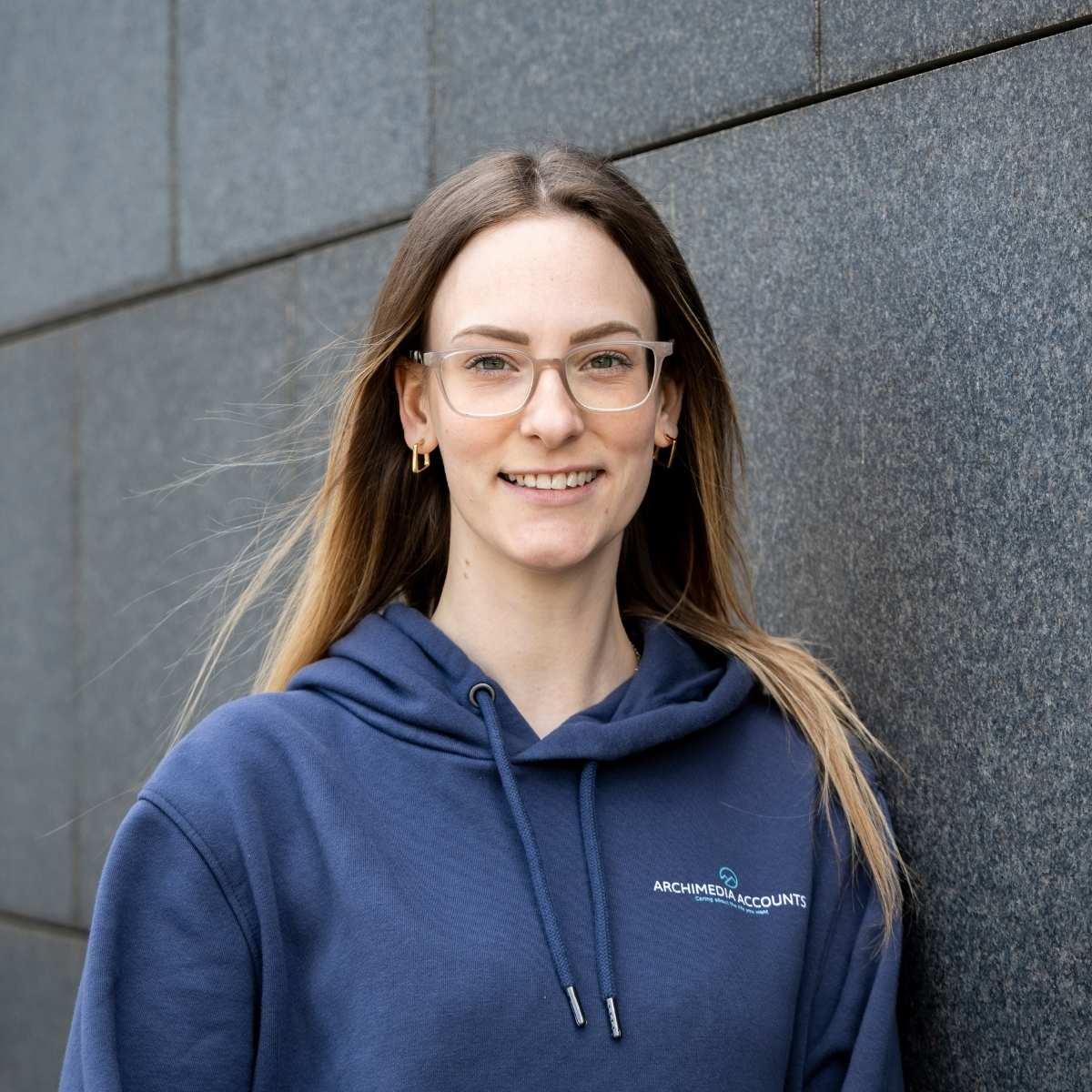 Team Photos - Archimedia Accounts - individual headshot in front of a dark grey wall wearing a branded hoodie