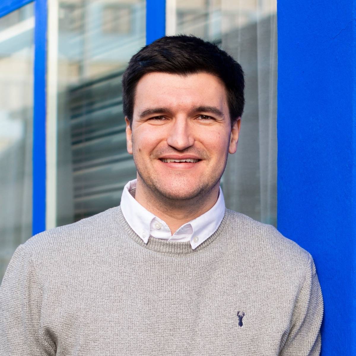 Headshot of Alex Pycock. Alex leans into a bright blue window frame. He wears a white collared shirt with grey jumper over the top, and smiles into the camera.