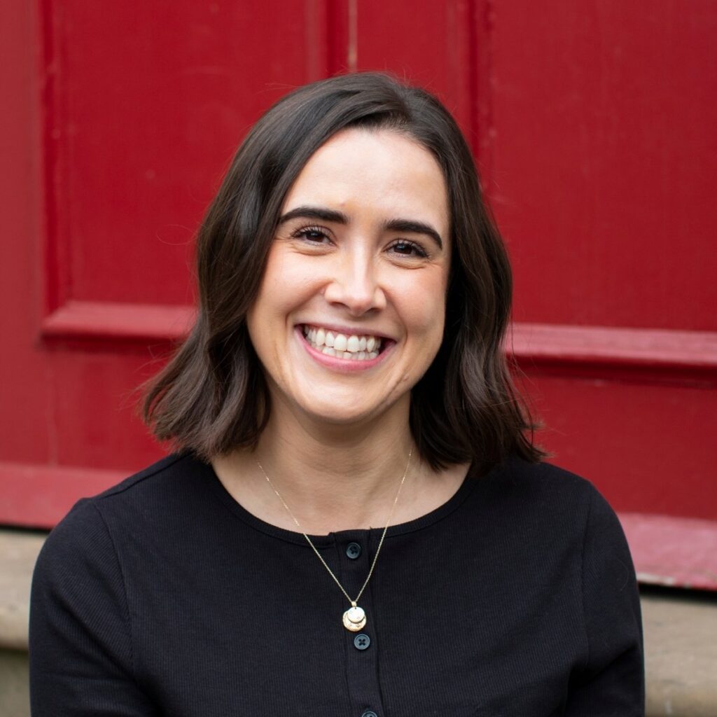 Headshot of Chloe Holmes. Chloe sits in front of a bright red door. She's wearing a black top and gold necklace and is smiling towards the camera.