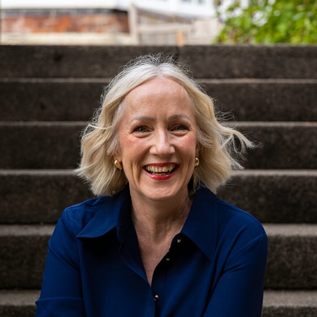 Headshot of Jen Stanbrook. Jen sits on some outdoor steps and wears a navy shirt and red lipstick. She's smiling towards the camera.